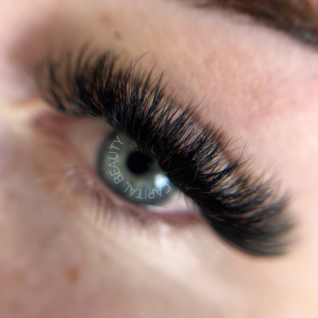 Close-up of an eye with long, thick, and curled eyelash extensions. The pupil reflects the words CAPITAL BEAUTY. The skin around the eye is smooth and slightly out of focus.