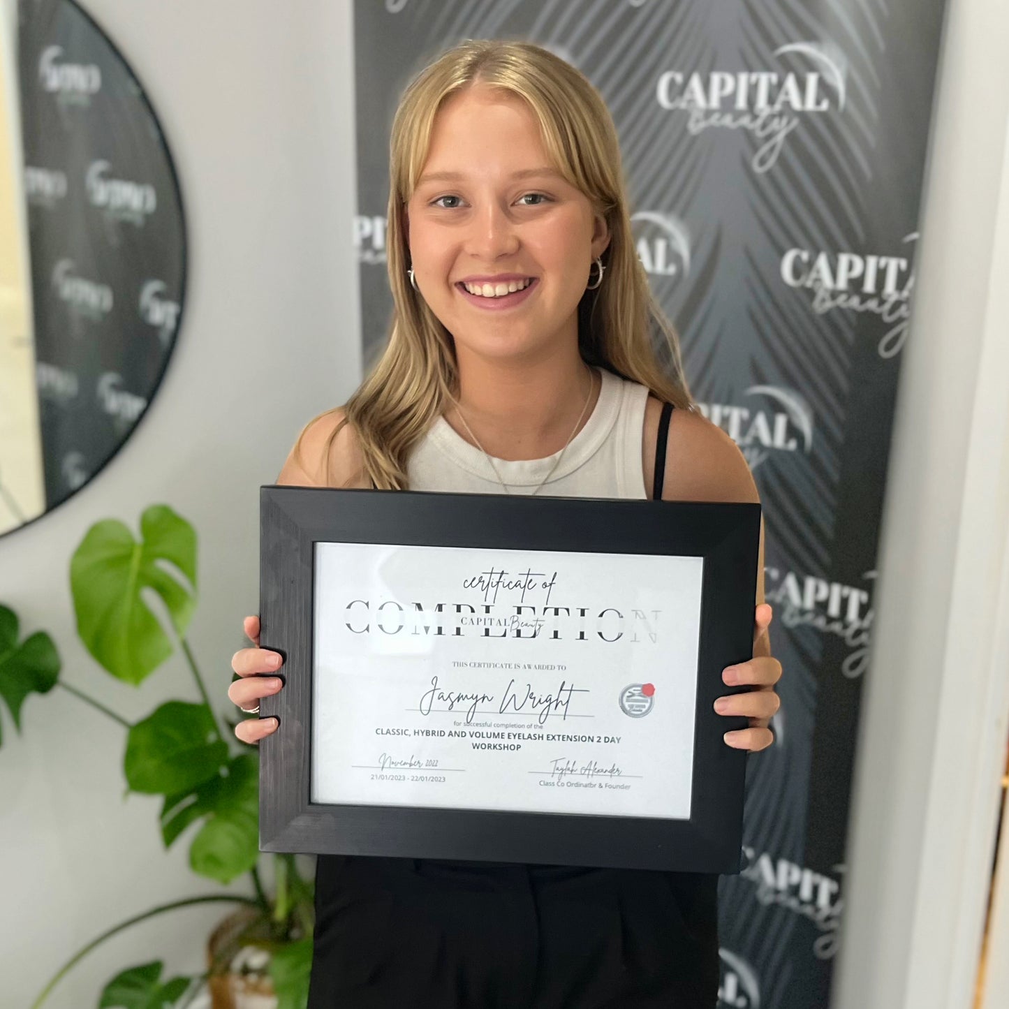 A smiling young woman stands indoors, holding a framed certificate of completion. Behind her is a banner with the words CAPITAL LASHES and a green potted plant is visible to the side.