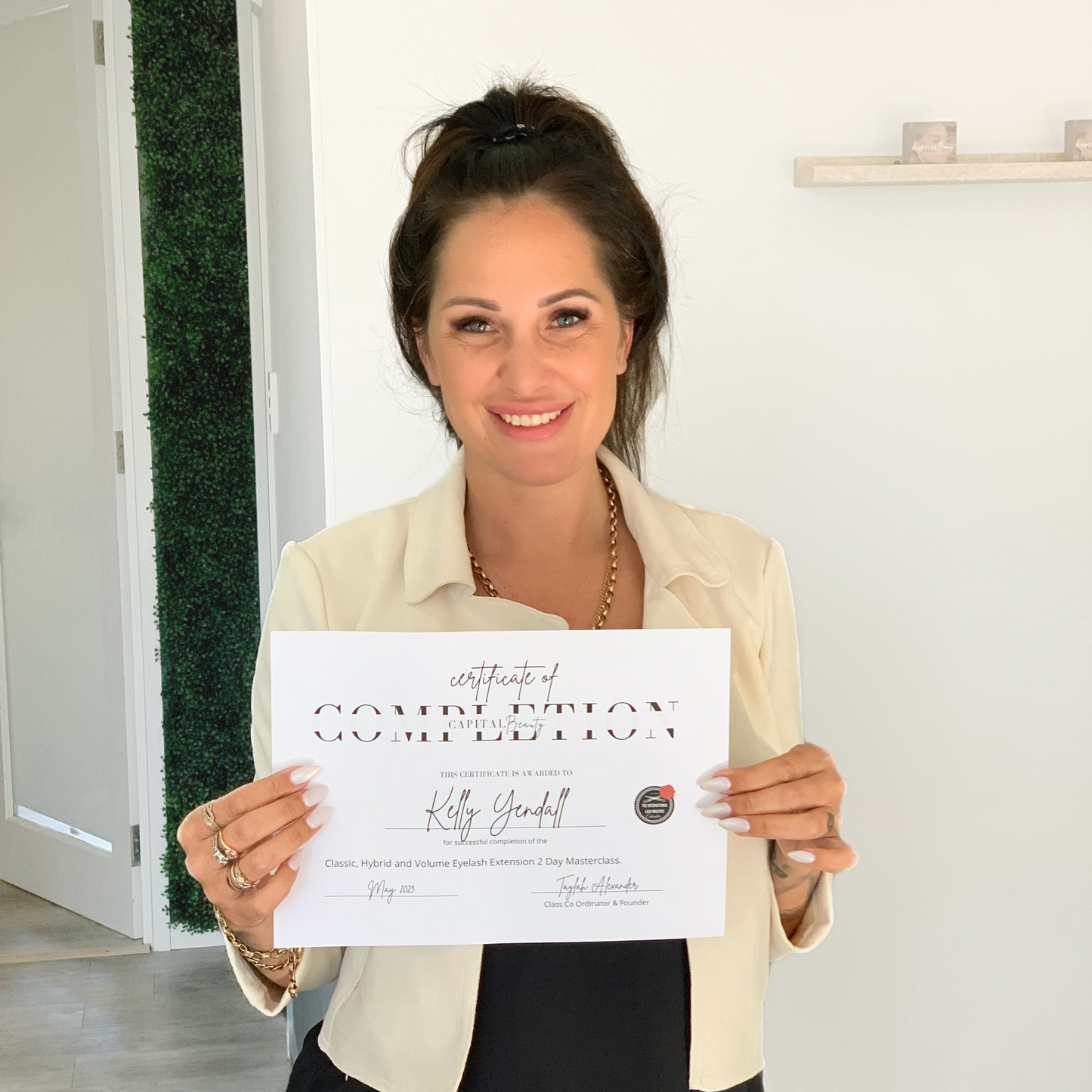 A woman with dark hair in a bun, wearing a white blazer, smiles and holds up a certificate of completion in a bright, modern room.