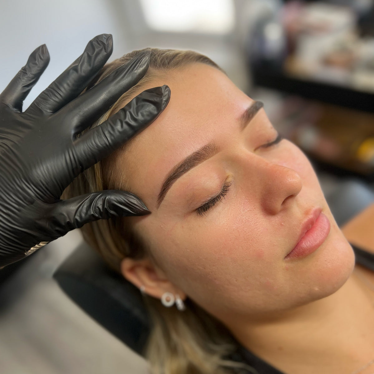 A woman with closed eyes relaxes as a gloved hand gently touches her forehead, highlighting her freshly shaped eyebrows.