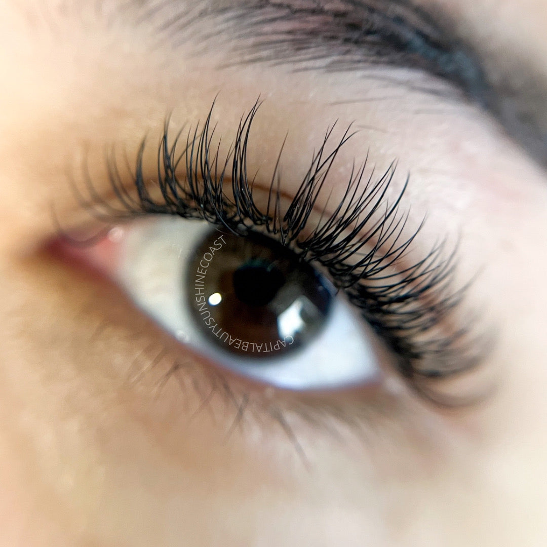 A close-up of an eye with long, thick, and curled eyelash extensions. The skin around the eye is smooth, and the lashes appear dark and voluminous.