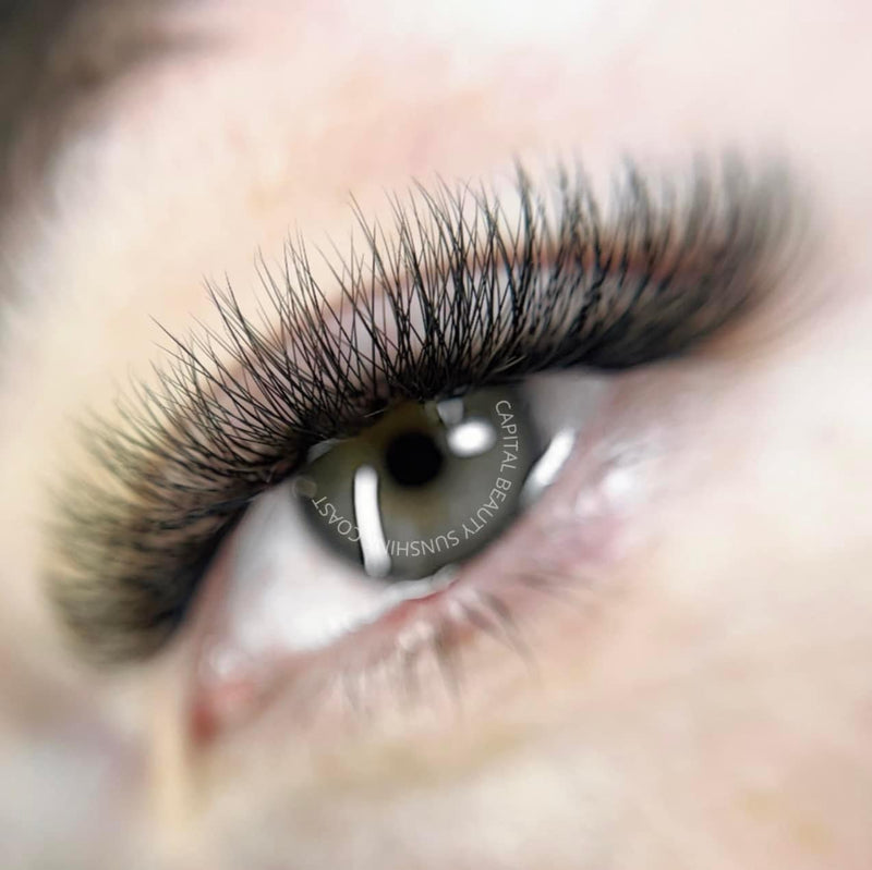 A close-up of an eye with long, thick, and curled eyelash extensions. The skin around the eye is smooth, and the details of the eyelashes are sharply in focus.