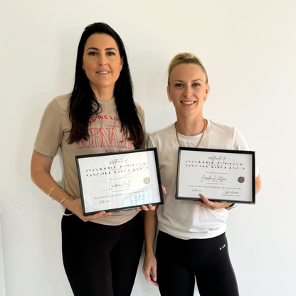 Two women stand side by side against a white wall, smiling and holding framed certificates of completion. Both are wearing casual clothes, including t-shirts and leggings.