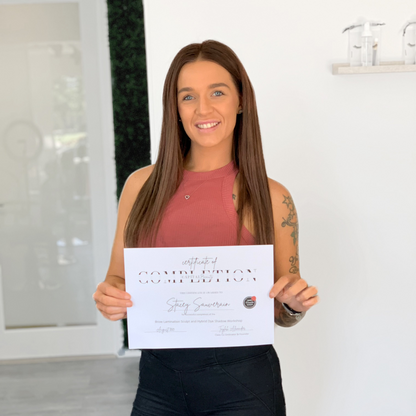 A woman with long brown hair, wearing a sleeveless mauve top and black pants, stands indoors smiling and holding a certificate of completion.