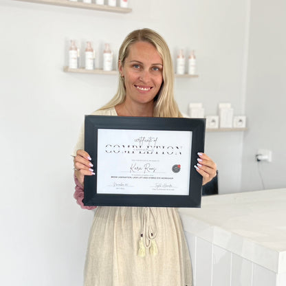 A woman with long blonde hair smiles while holding a framed certificate of completion in front of a white counter and shelves with skincare products in a bright, modern room.