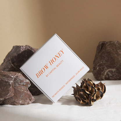 A white box labeled Brow Honey by Capital Beauty stands upright among brown rocks and dried plant decor, with text highlighting No White Residue, Waterproof, 24 Hr Hold.