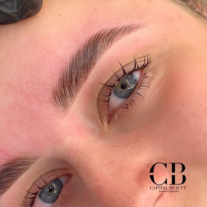 Close-up of a person’s face showing freshly shaped and tinted eyebrows and long, curled eyelashes. The skin appears smooth and the brow area is defined. The Capital Beauty logo is visible in the lower right corner.